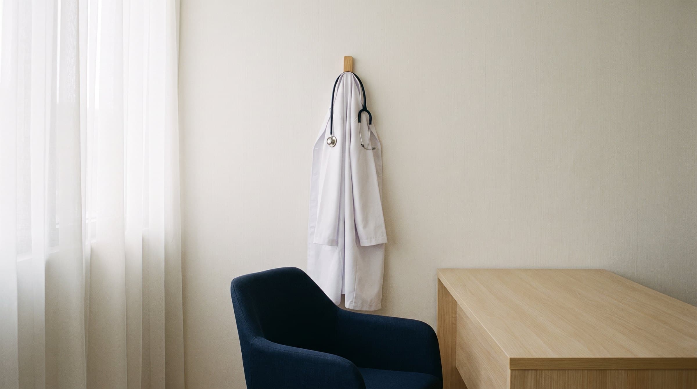 A doctor's white coat and stethoscope on a hook in an empty consultation room
