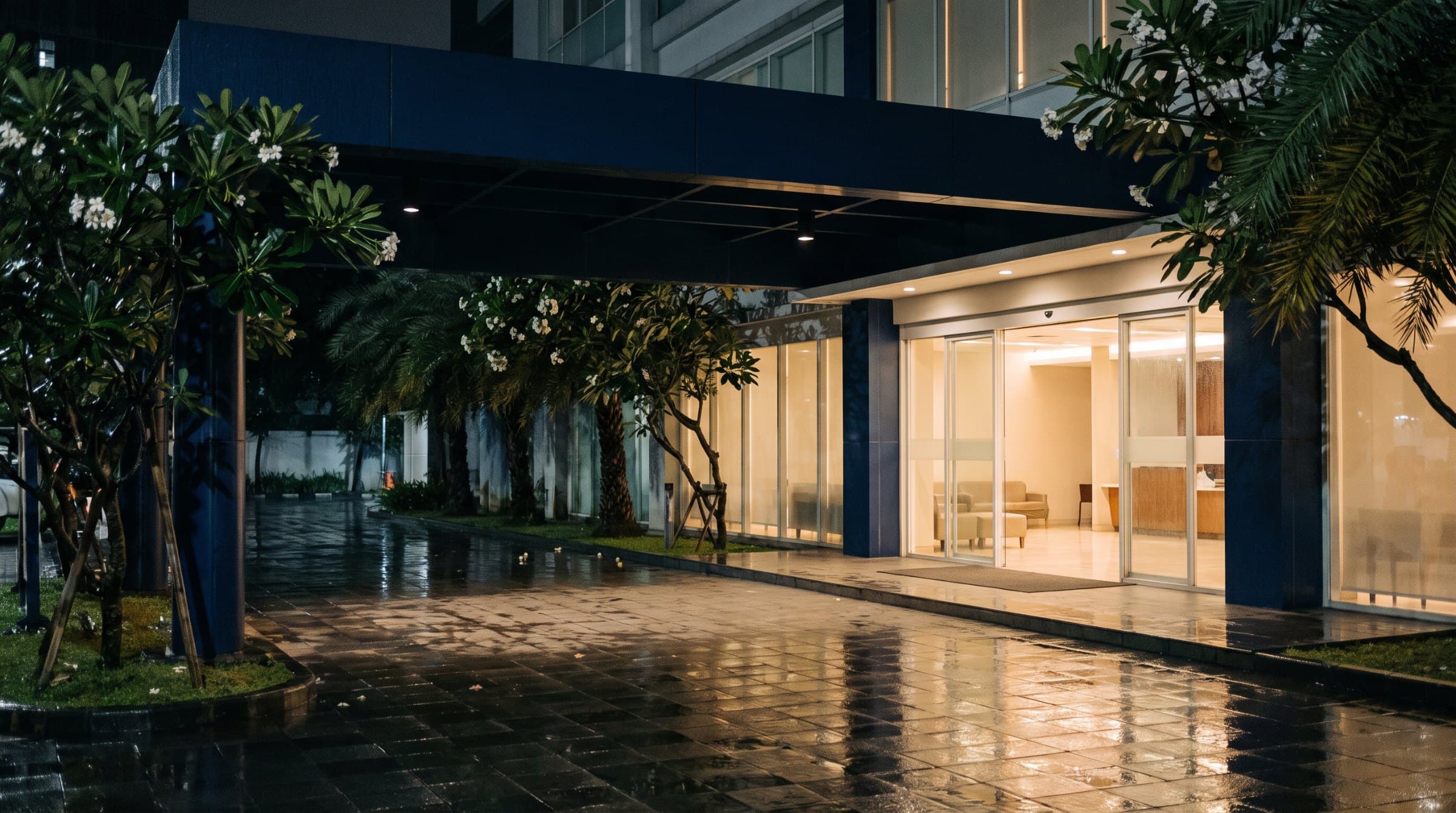 A hospital entrance at night after rain, its glass doors lit from the lobby inside